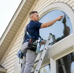 Professional window cleaner on a ladder using a squeegee to clean an exterior arched window, Salt Lake City, UT.